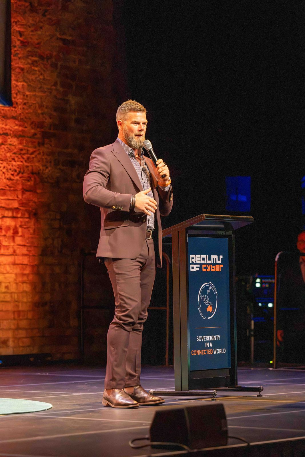 Adam Green speaking into a handheld microphone beside the Realms of Cyber podium, a lit brick wall behind him.