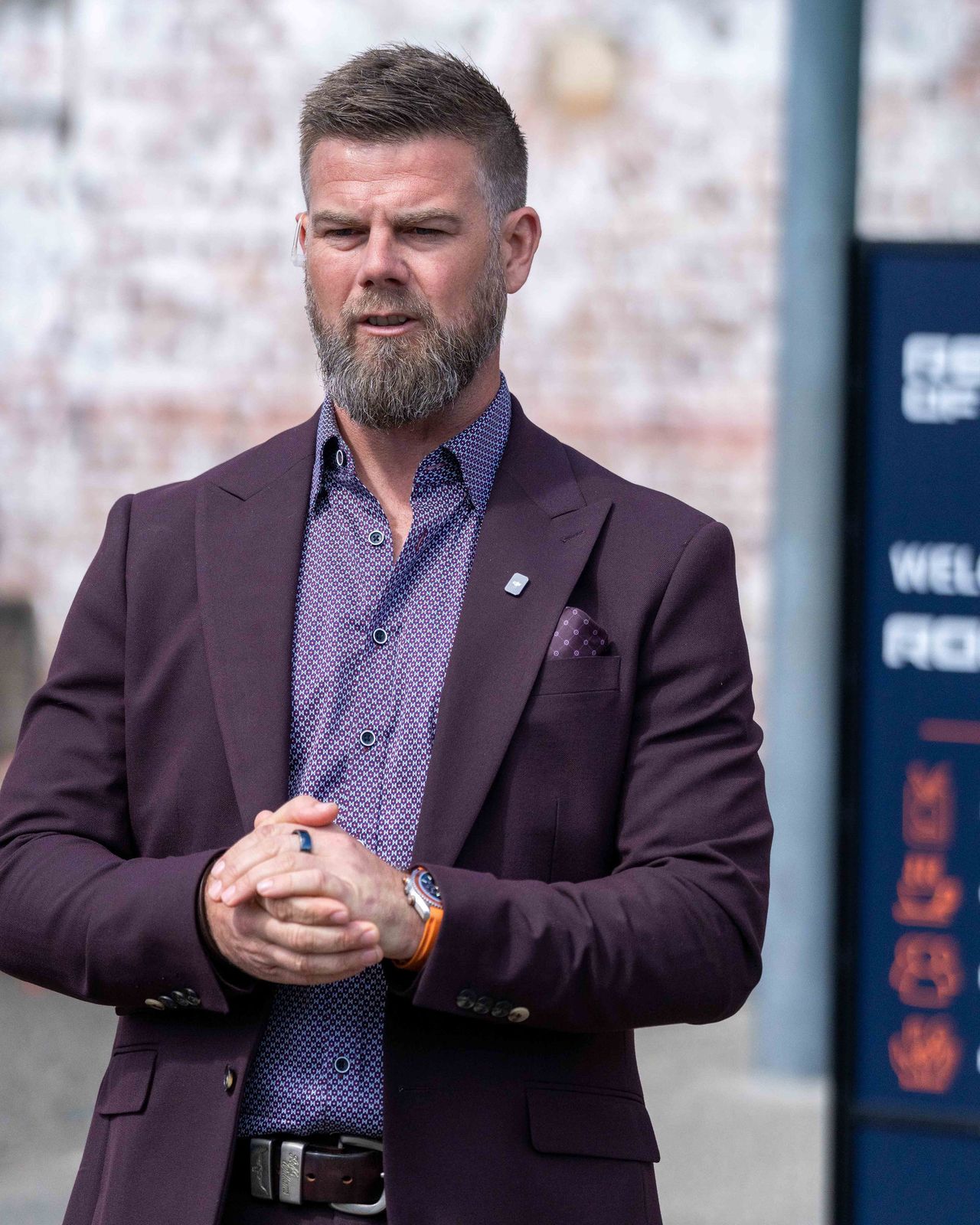 Adam Green standing outdoors in a plum suit, a clear earpiece tube behind his ear.